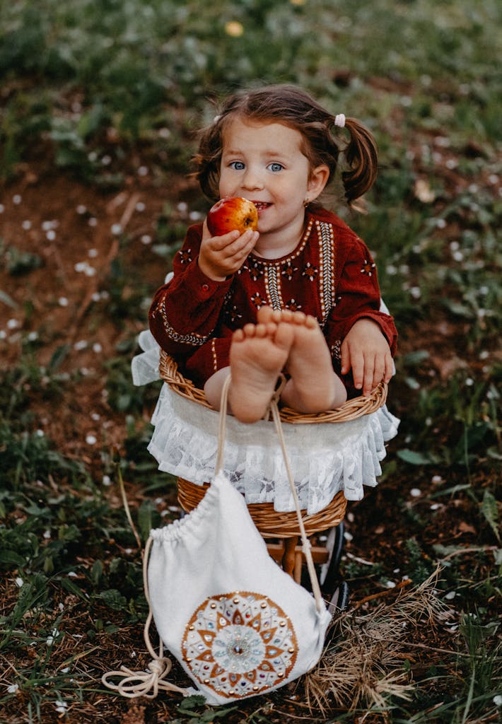 Cute child with pigtails sitting in a basket outdoors, enjoying an apple in Slovakia.