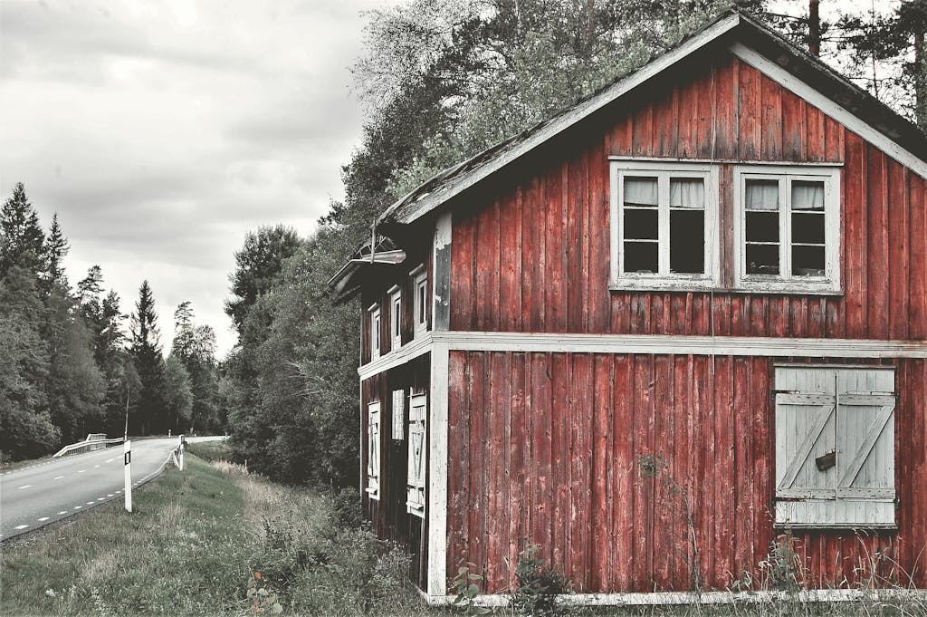Old red wooden farmhouse along a rural forest road under cloudy skies, exuding rustic charm.